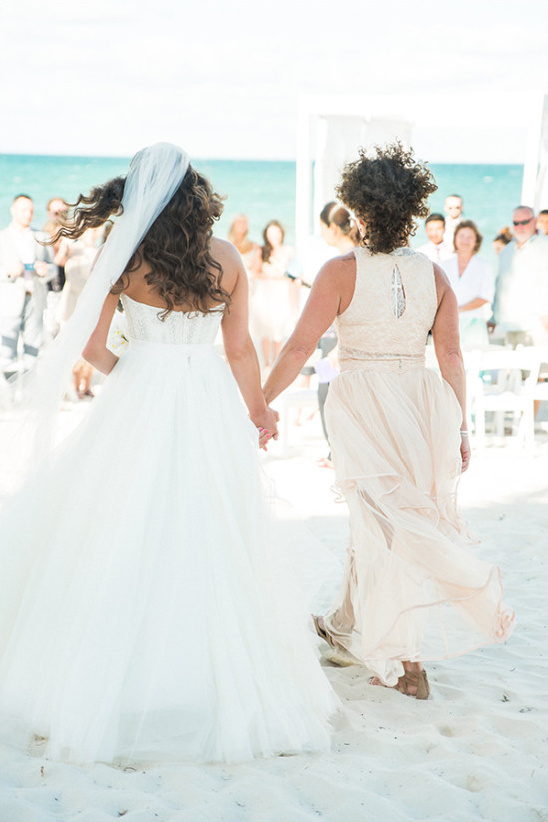 Bride and mother walk hand in hand at a beach wedding ceremony