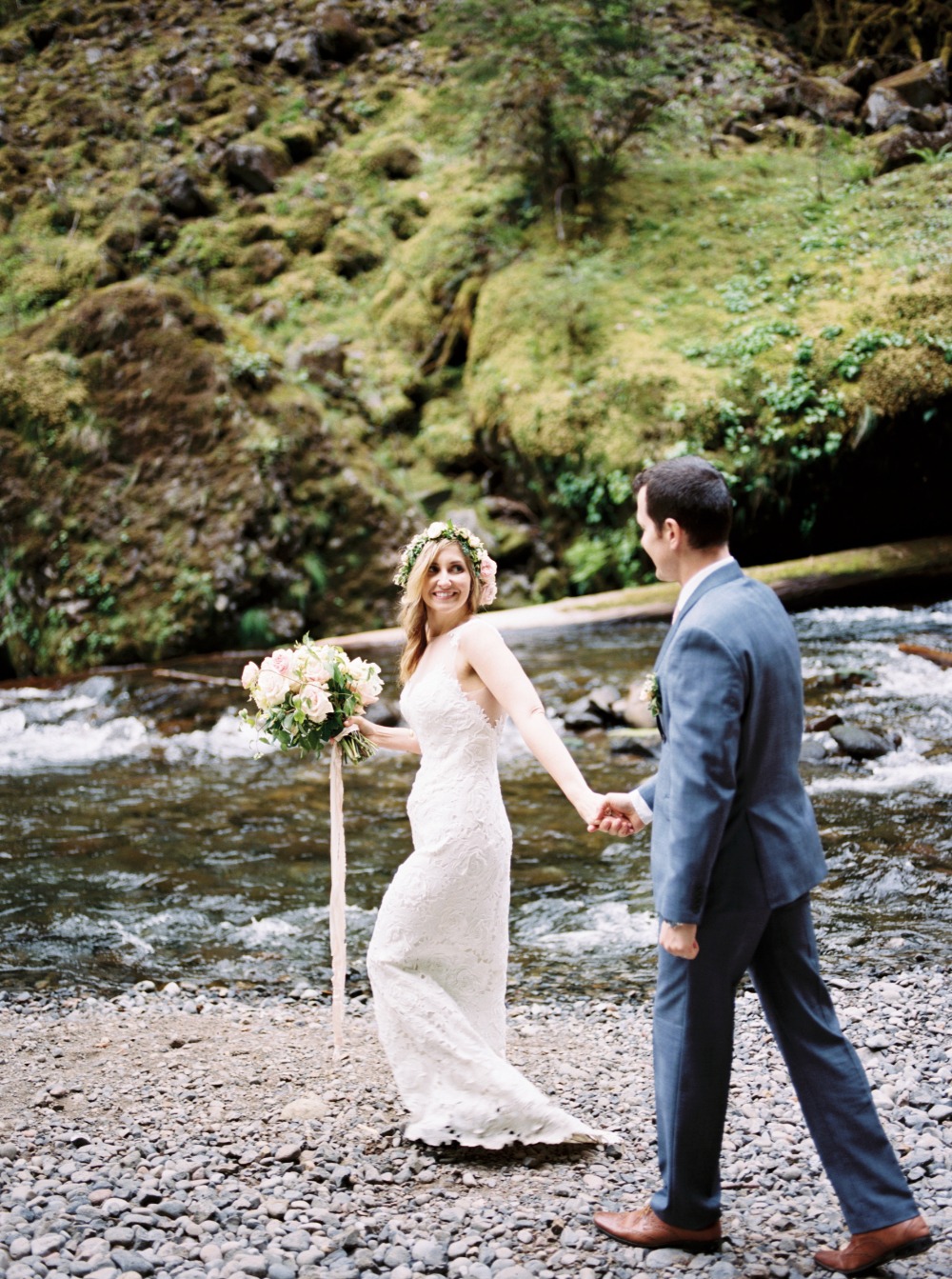 Bride and groom hold hands by a scenic river on wedding day