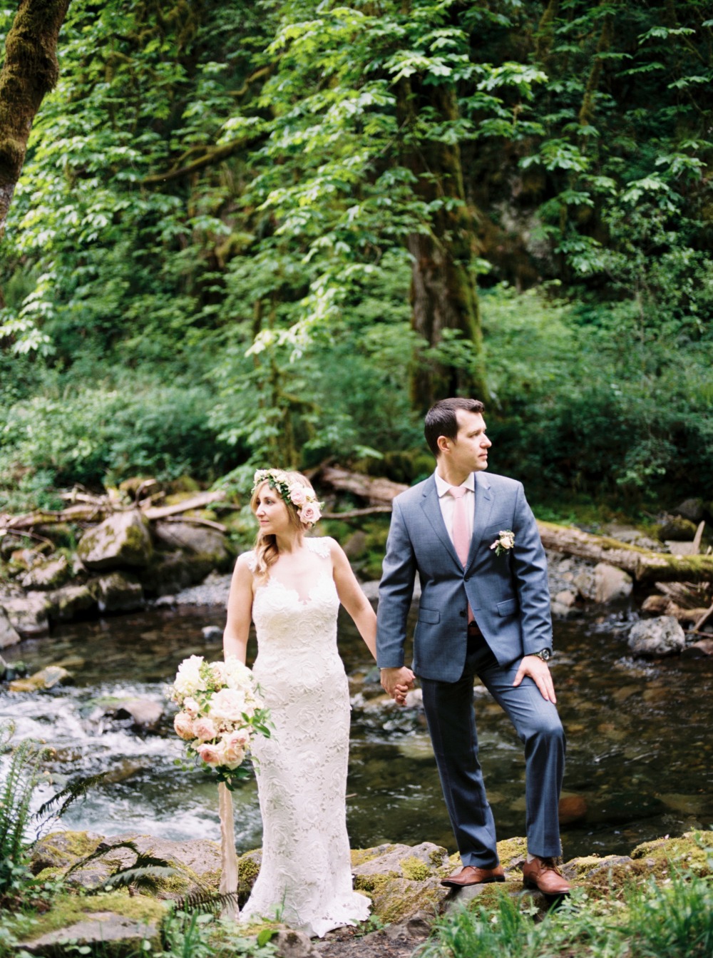 Bride and groom hold hands by creek in forest wedding