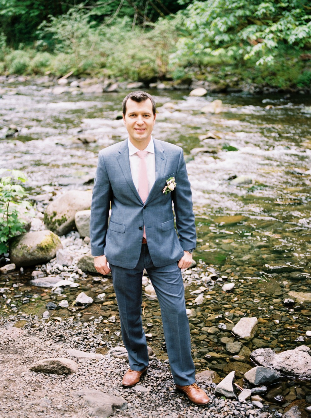 Groom in suit stands by rocky river on wedding day