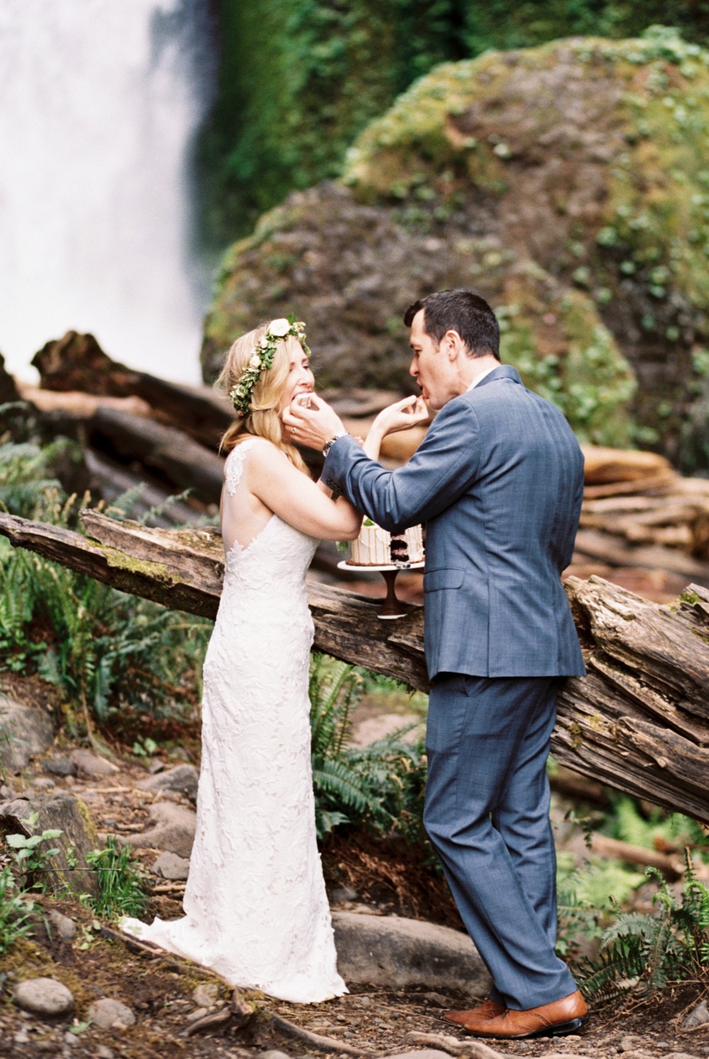 Bride and groom share wedding cake by waterfall