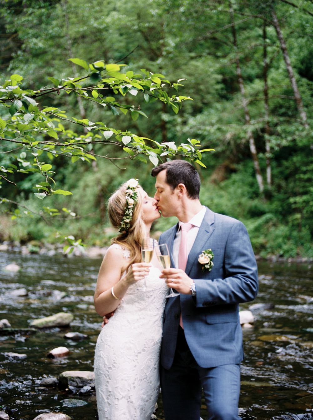 Bride and groom kissing by river, toasting with champagne