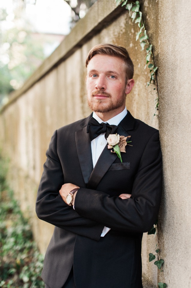 Groom wearing a black tuxedo leaning against a wall, ready for his wedding