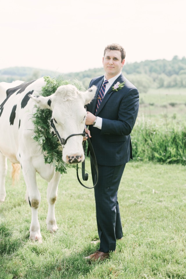 Groom poses with a cow wearing a floral wreath on a beautiful meadow