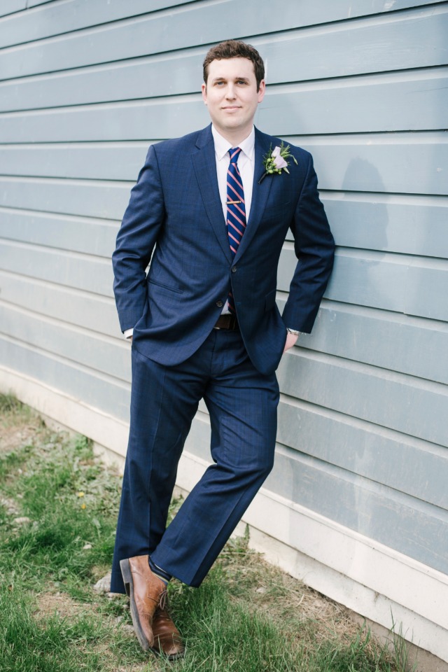 Groom in blue suit leans against blue wall outdoors