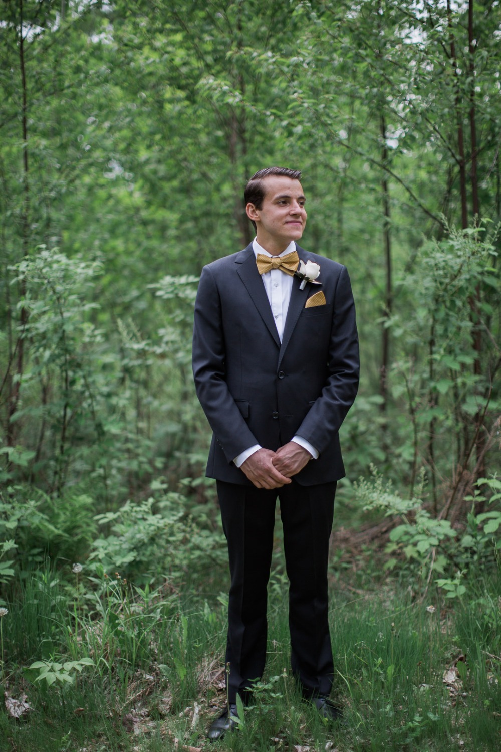 Groom in forest, wearing dark suit and gold bow tie