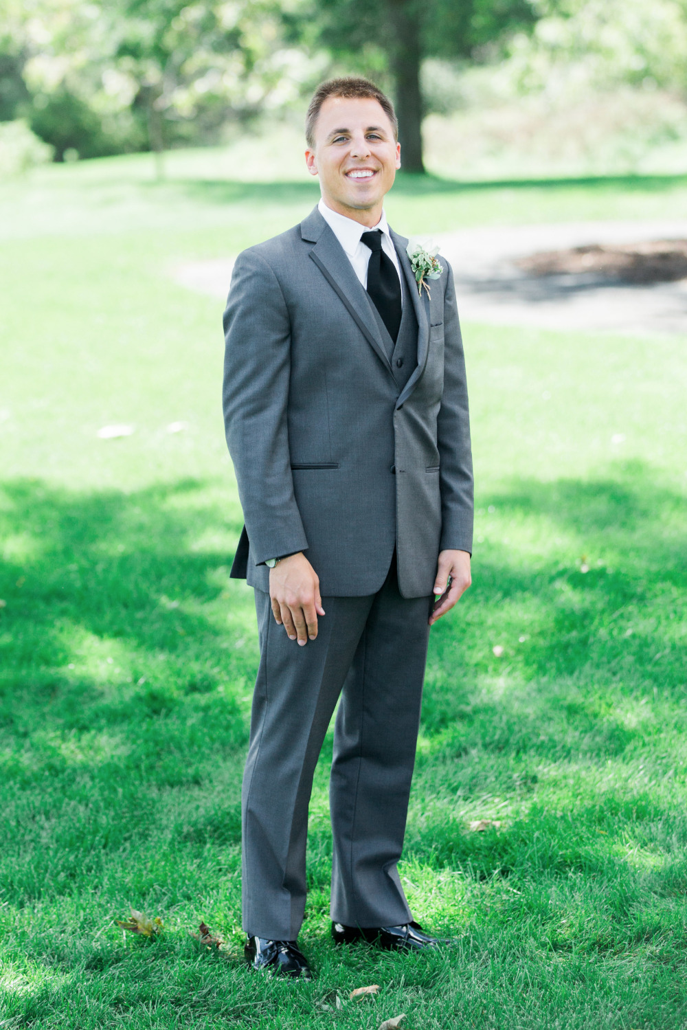 Groom smiles in a gray suit and black tie on grass
