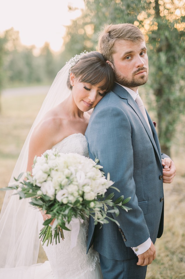 Bride leans on groom at a wedding, holding white flowers