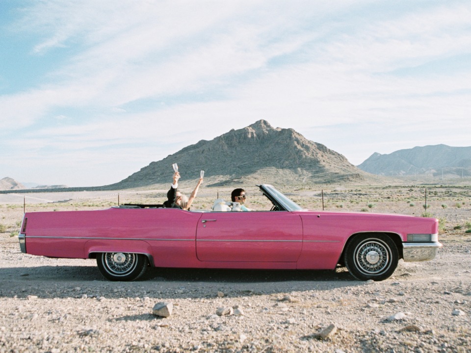 People in a pink convertible toast in the desert with a mountain