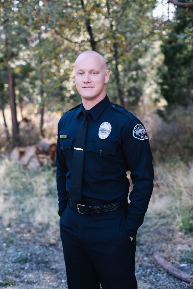 A bald man in a police uniform stands outdoors and smiles at camera