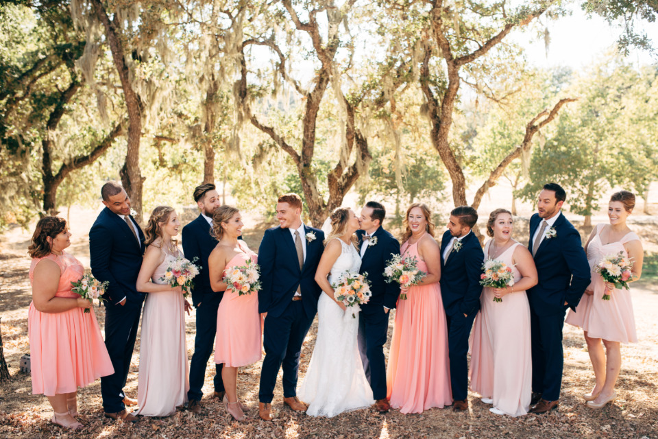 Wedding party under oak trees, bride and groom kissing