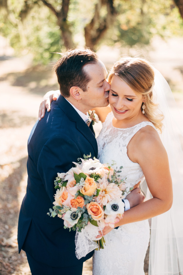 Happy bride and groom share a kiss, holding peach bouquet