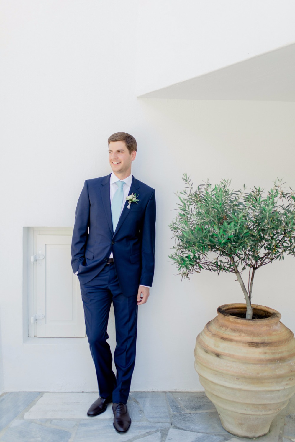 Groom in navy suit smiles beside a decorative olive tree