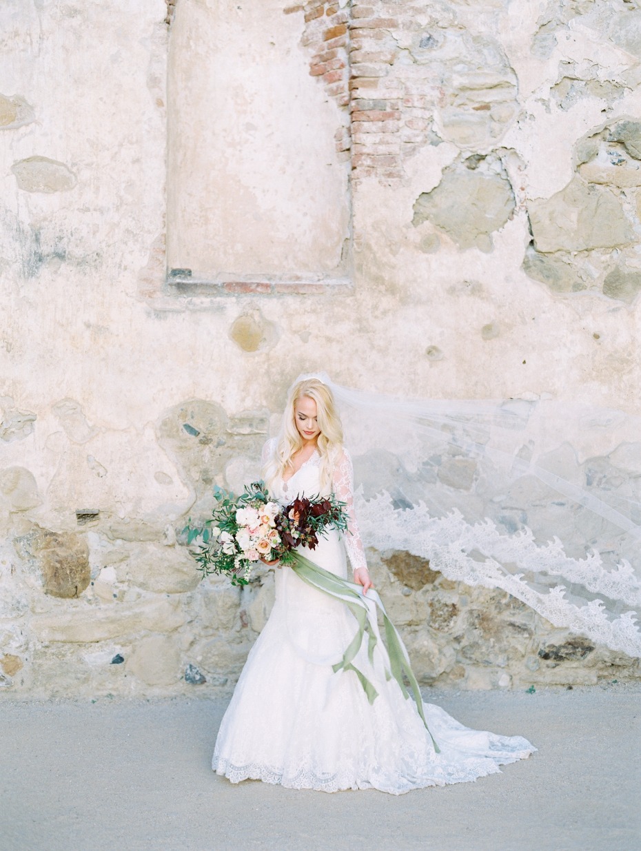Bride in lace gown holding a bouquet against stone wall