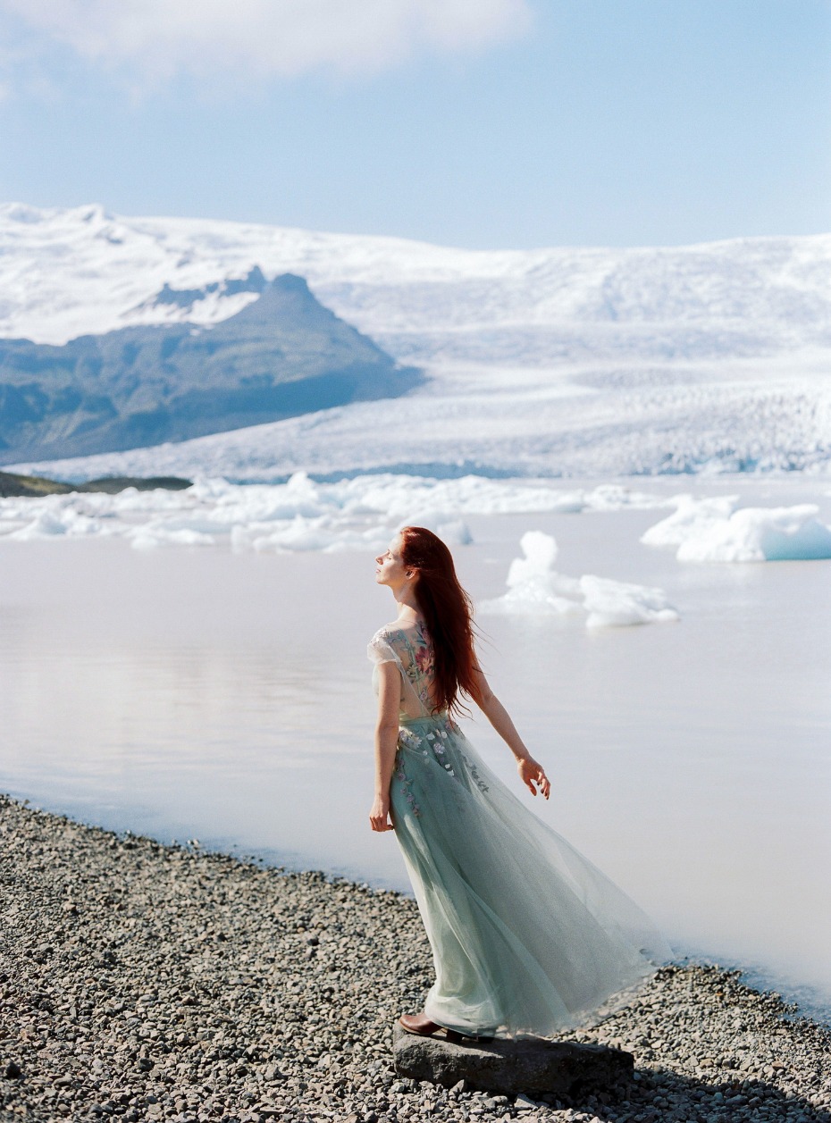 Woman in a flowing dress stands by a glacial lake, looking up