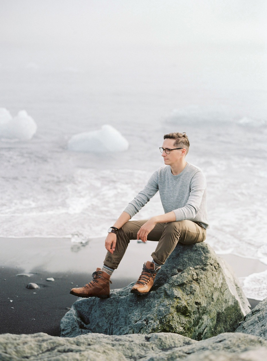 Man in glasses sits on a rock at the seaside, contemplating the ocean view