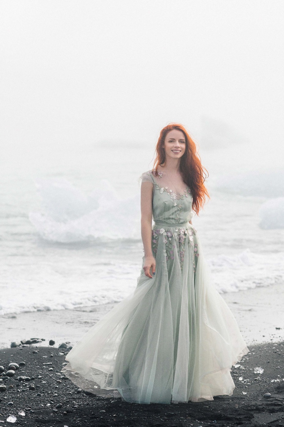 A woman in a flowing green dress smiles on a dark beach