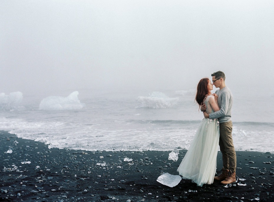 Couple embraces on a black sand beach with icebergs, foggy day