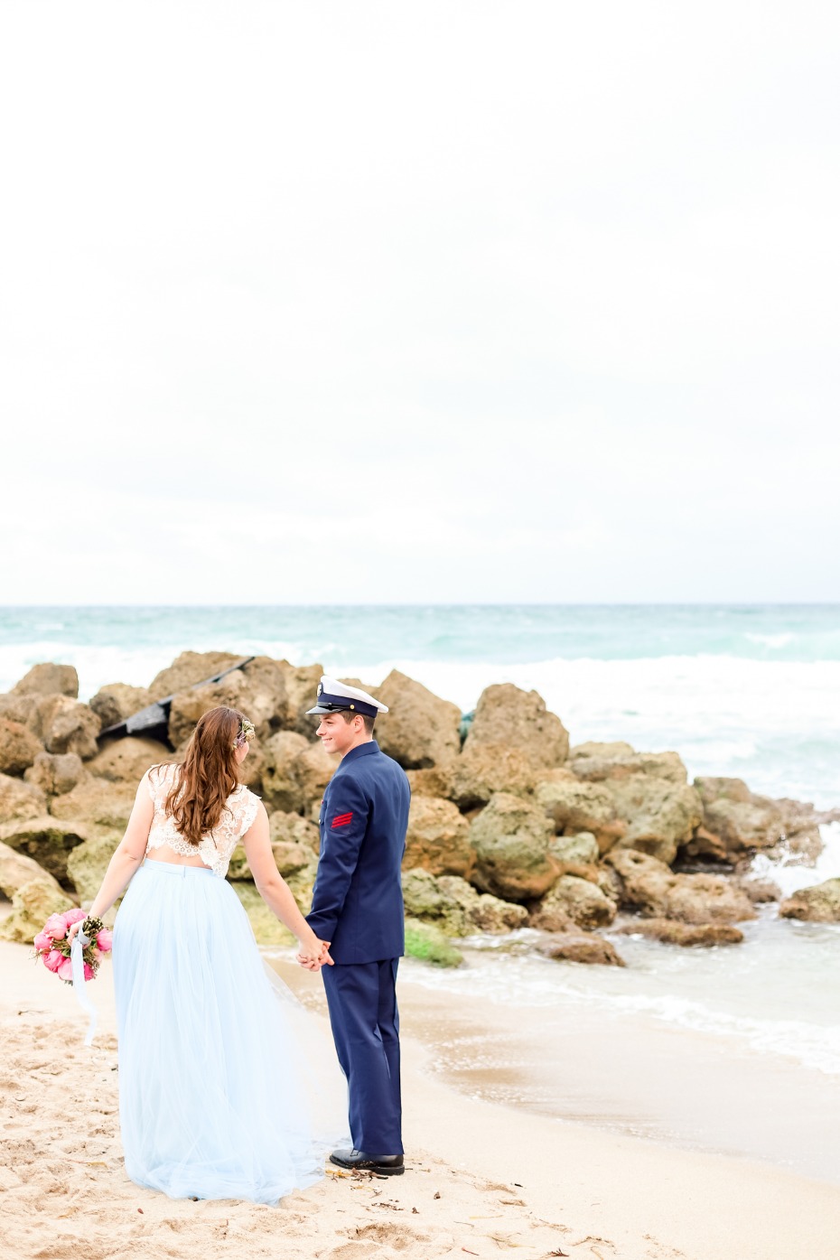 Bride and groom hold hands on a beach near the ocean