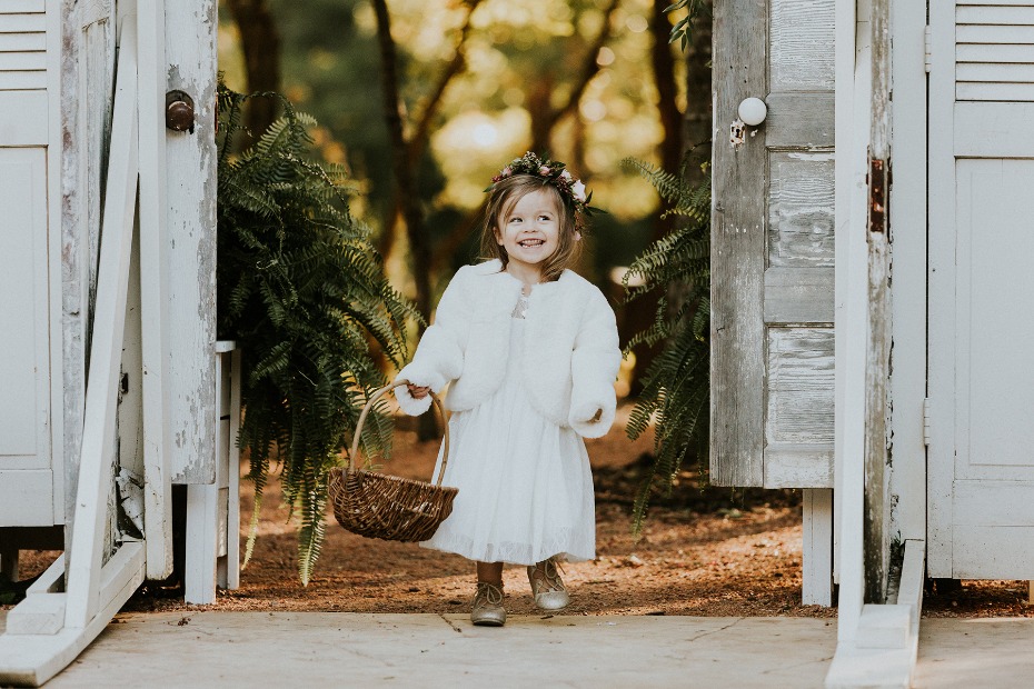 Smiling flower girl walks through rustic doorway with basket