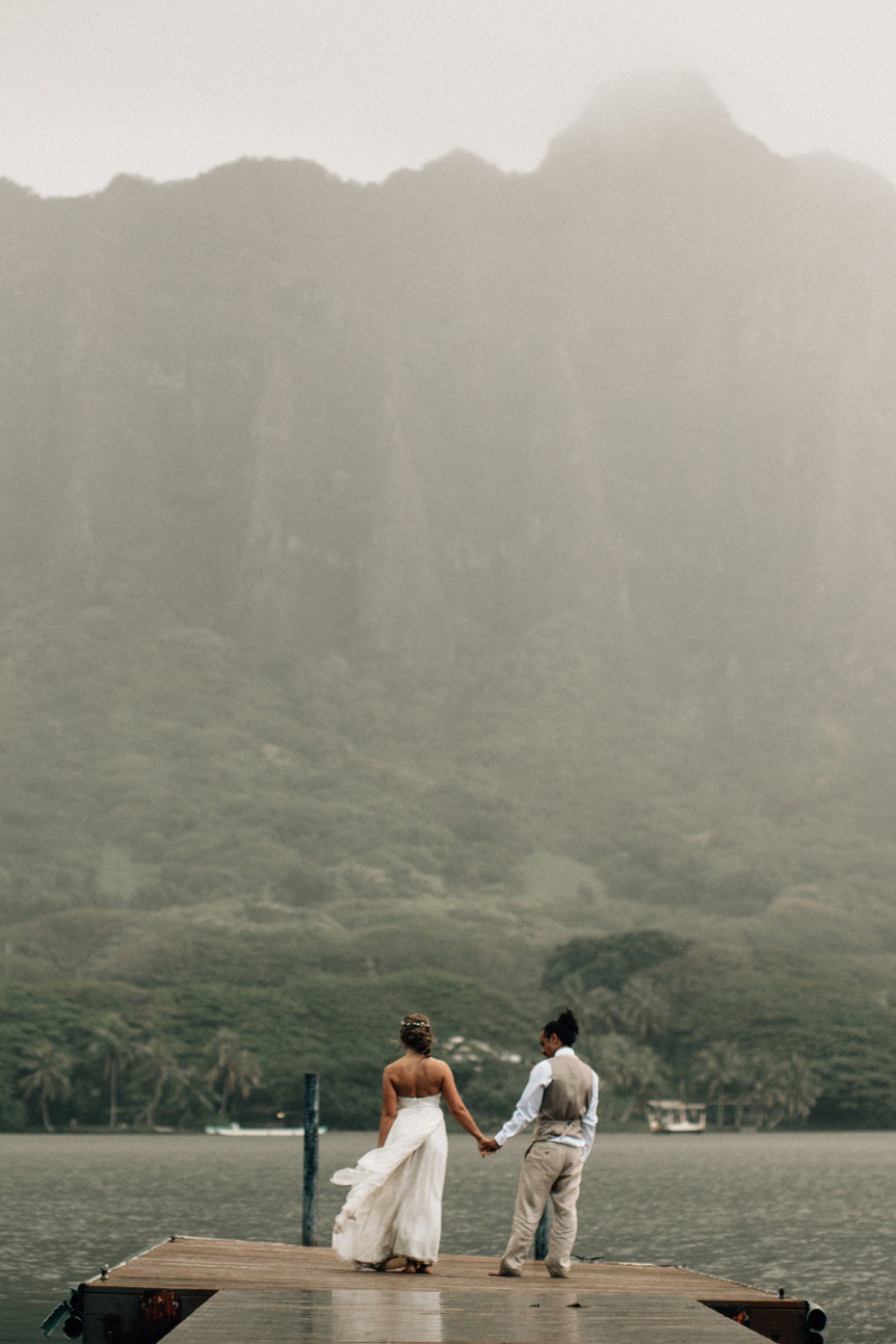 Newlyweds hold hands on dock facing misty mountains