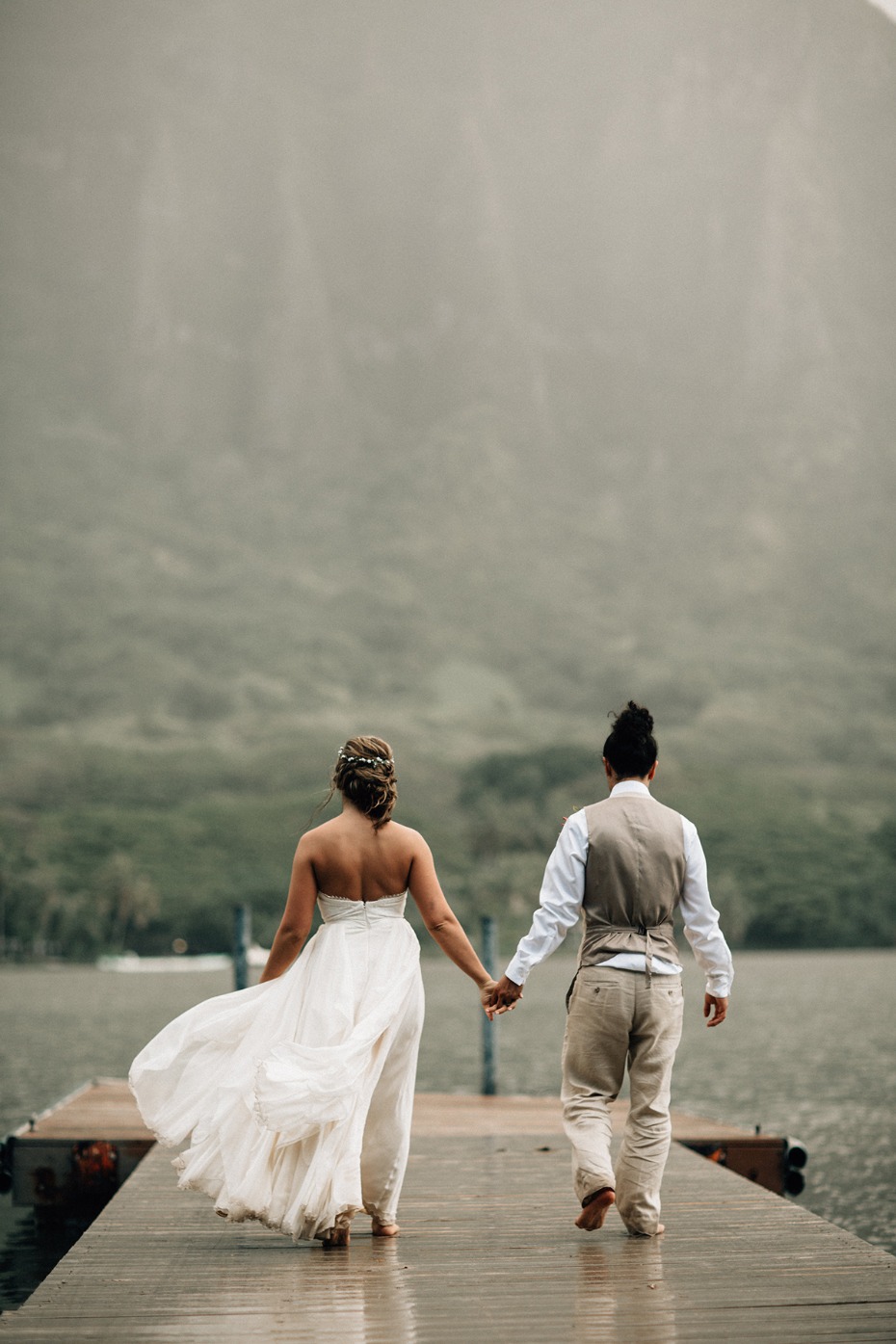 Newlyweds walk handinhand on a pier in misty mountains