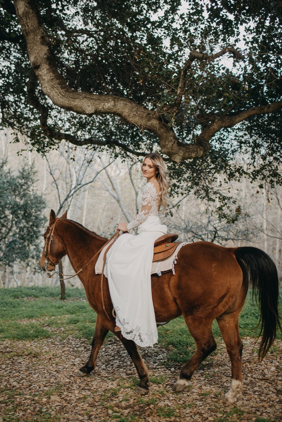 Bride in white gown riding a brown horse under a tree