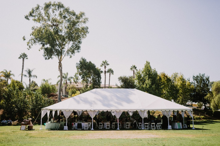 Outdoor event with a large white tent and surrounding trees on a sunny day
