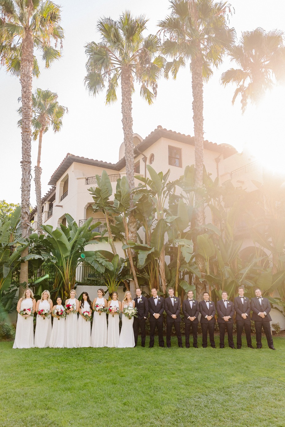 Wedding party poses in front of a mansion on green grass with palm trees