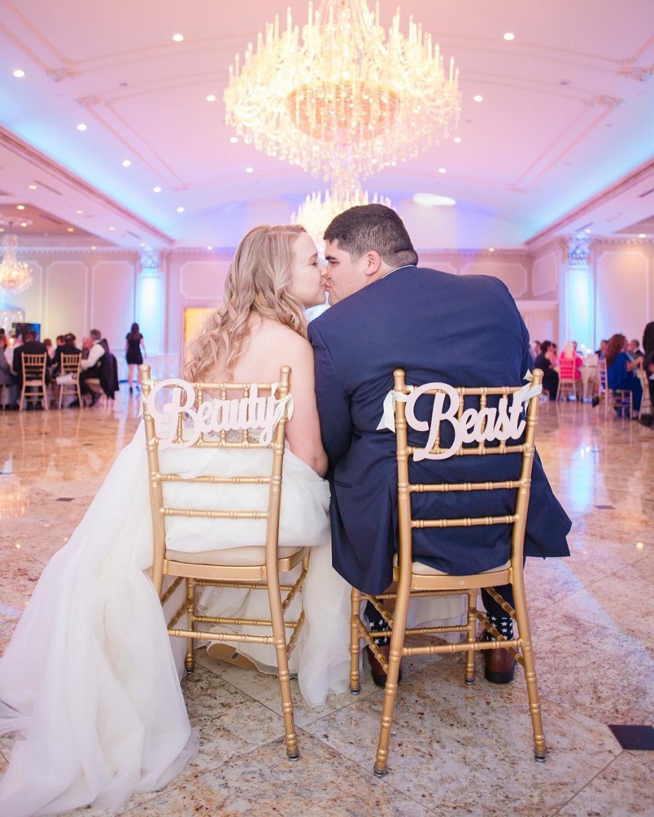 Bride and groom kissing at wedding reception, chairs say Beauty and Beast