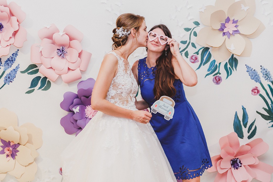 Bride and bridesmaid pose with props at a flowered wedding backdrop