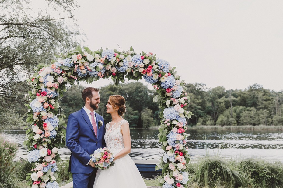 Couple under floral arch at wedding ceremony, lake in background