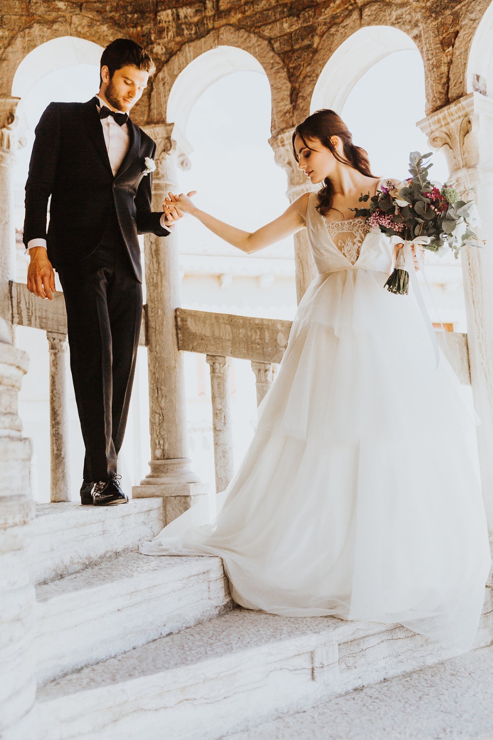 Groom gently guides bride down elegant stone stairs