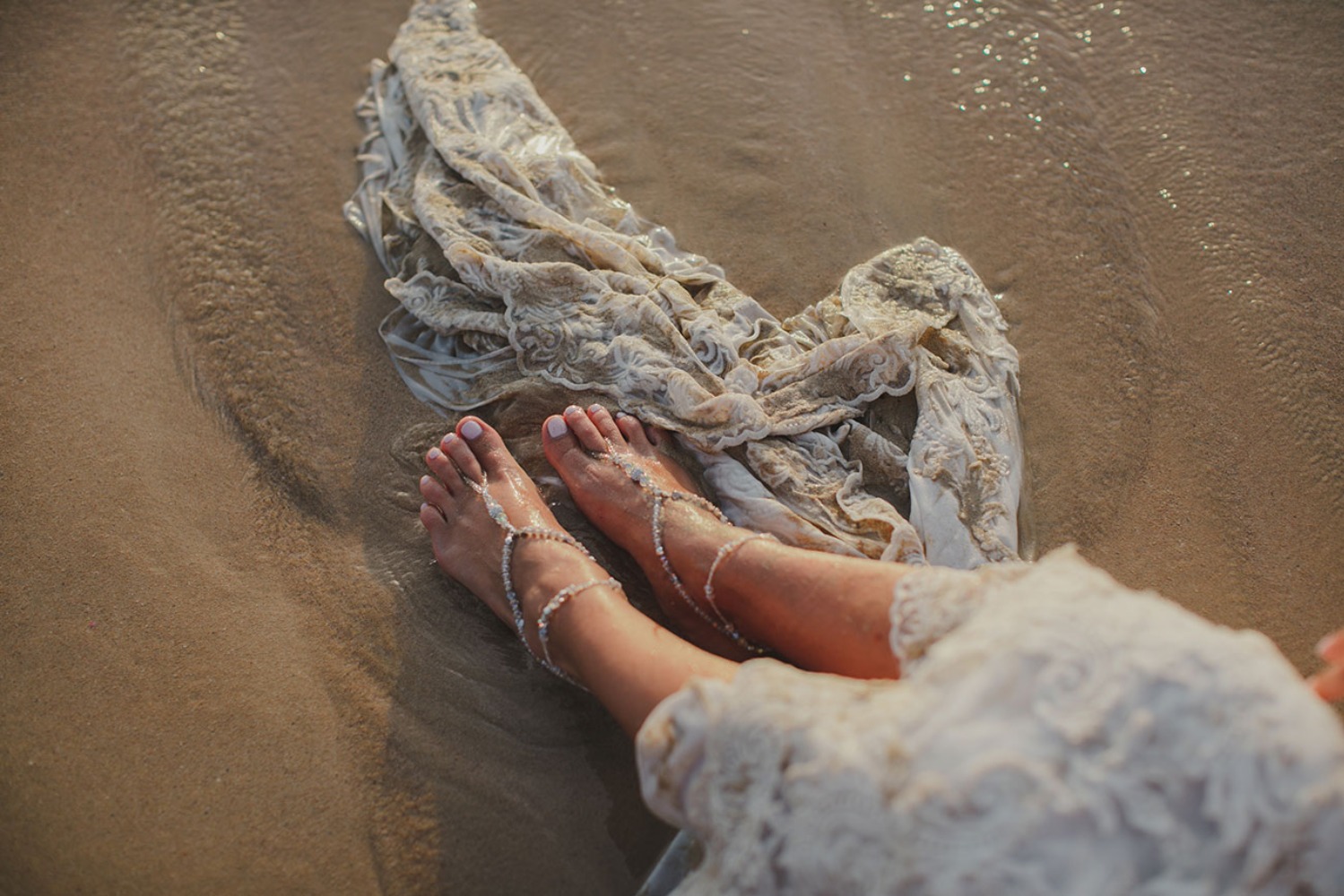 Feet with beaded anklets and wedding dress on sandy beach