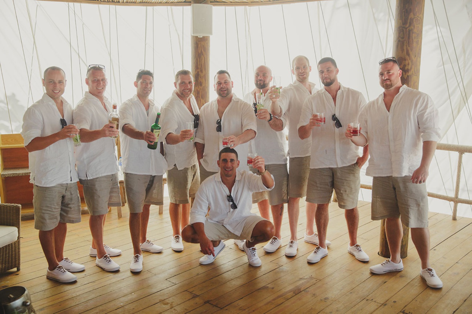 Groomsmen enjoying drinks in white shirts and shorts on wooden deck