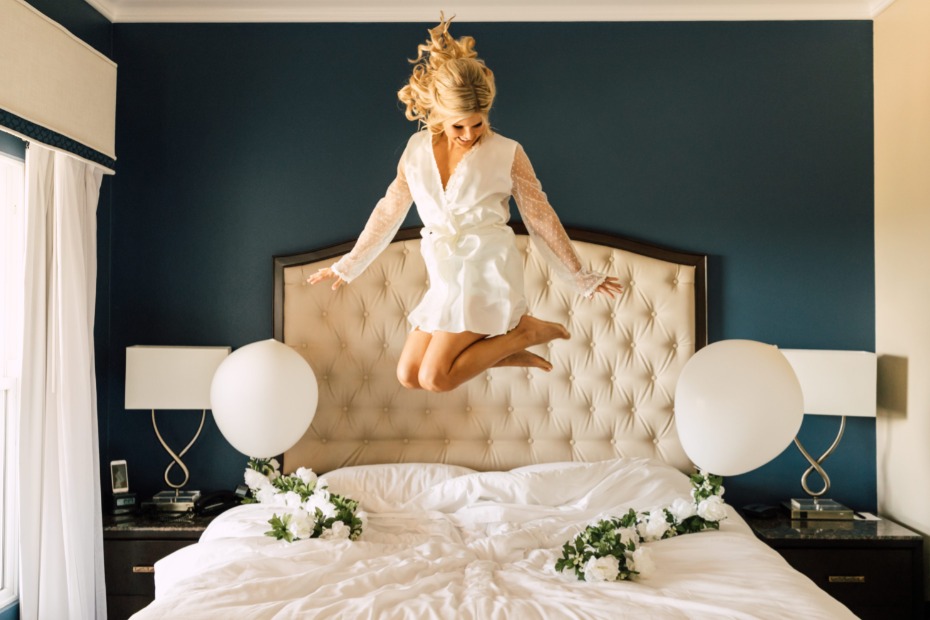 Bride in robe joyfully jumps on bed with floral accents