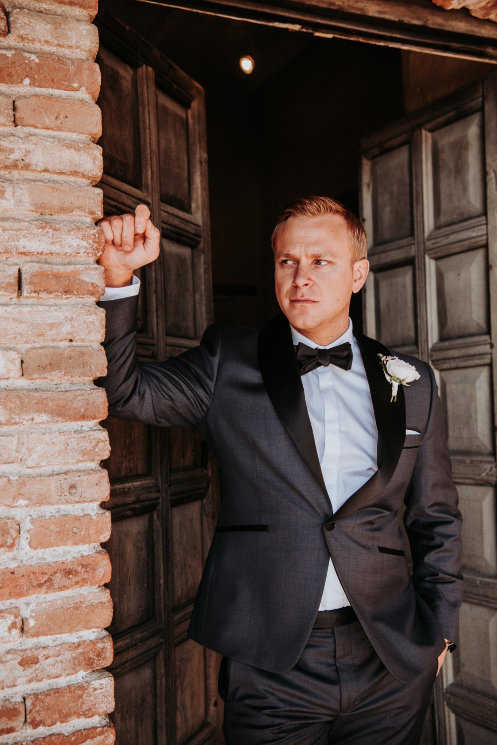 Groom in tuxedo poses by wooden doorway brick wall