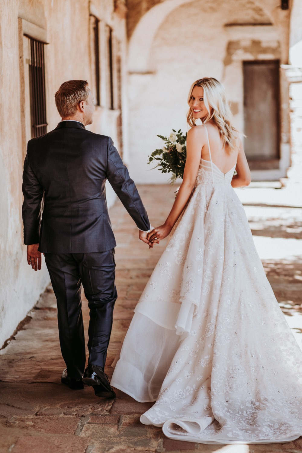Bride and groom walk, holding hands, through a historic building archway