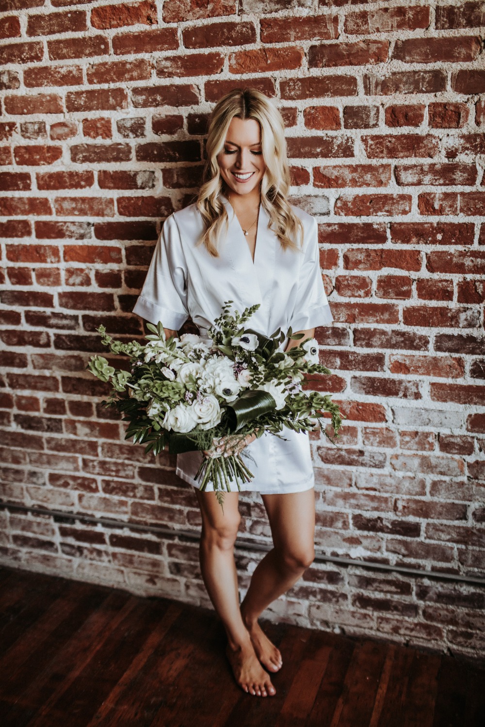 Bride in robe holding wedding bouquet near brick wall