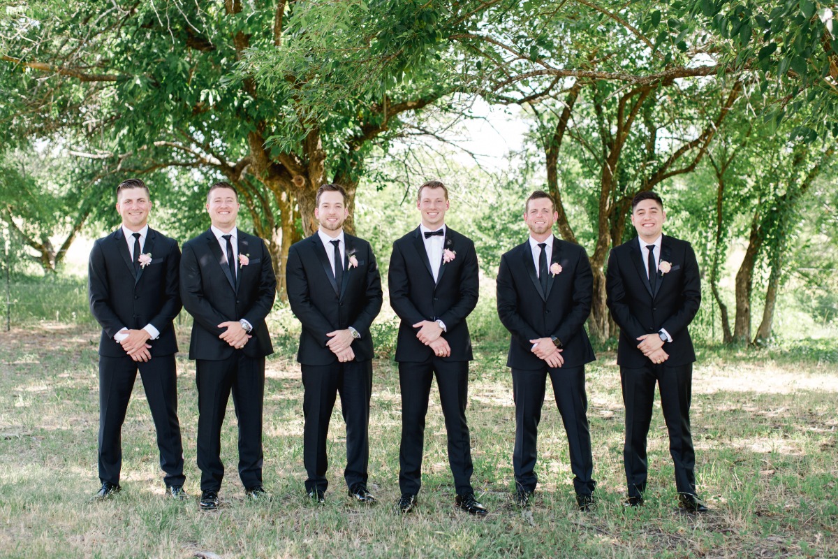 Groomsmen in black suits standing outdoors on a sunny day