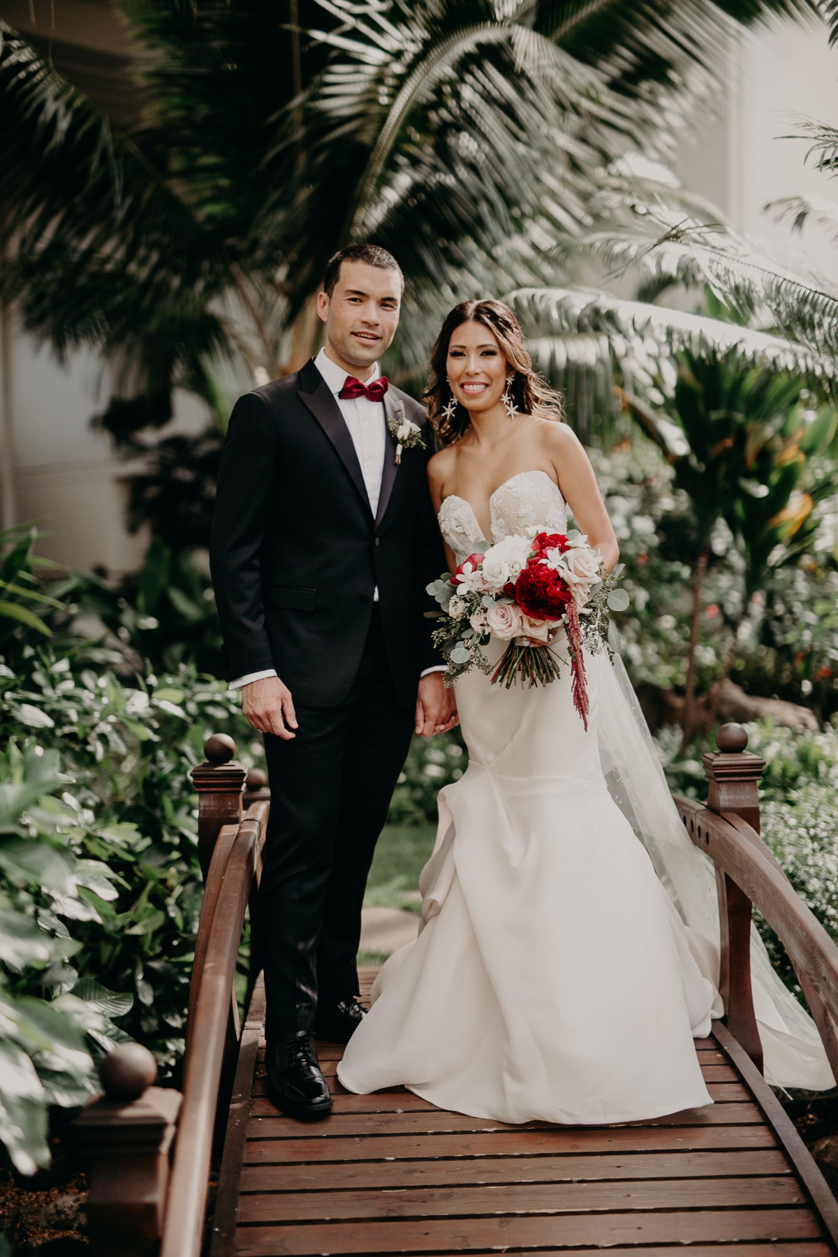 Bride and groom pose on wooden bridge, surrounded by greenery