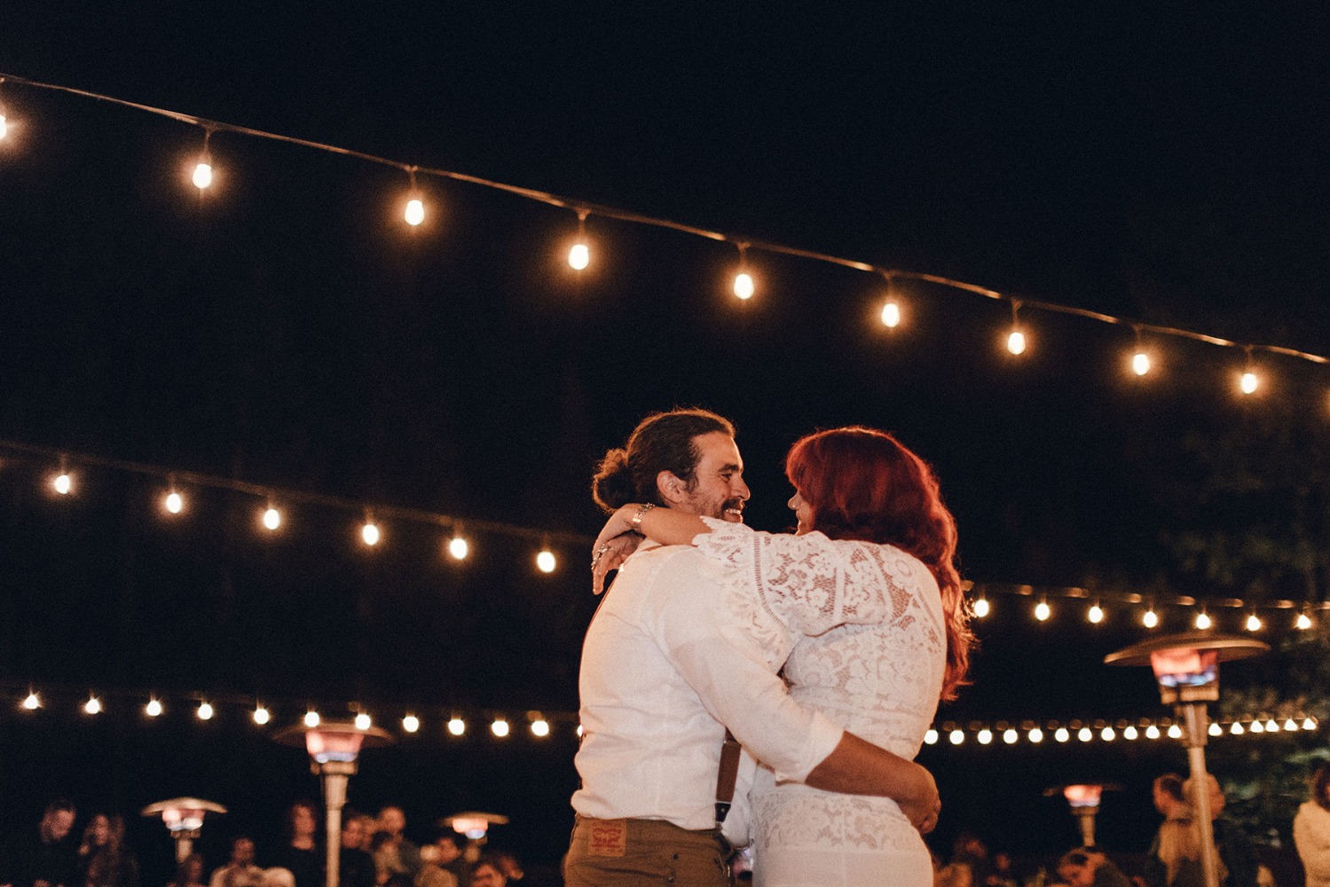 Newlyweds embrace on a dance floor under warm string lights at night