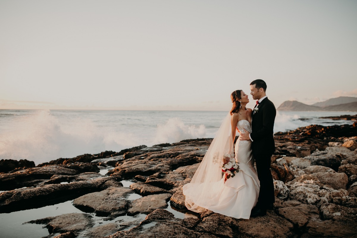 Elegant wedding couple embraces on a rocky shoreline at sunset