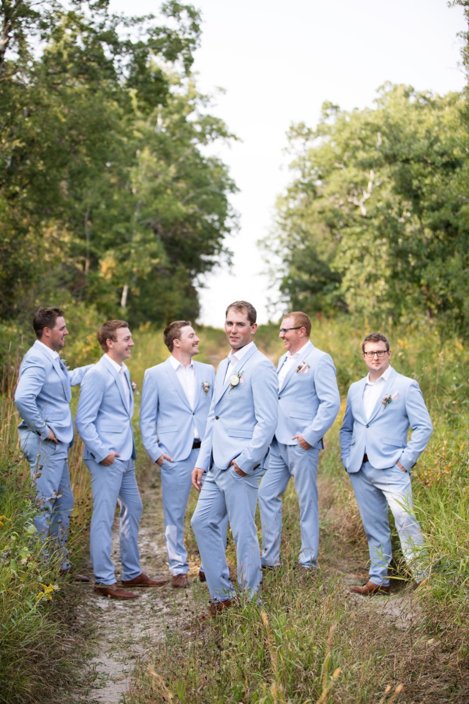Groomsmen wearing light blue suits stand on a path in nature