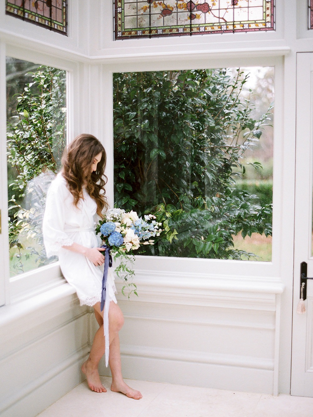 Bride in robe holds bouquet by window on wedding day