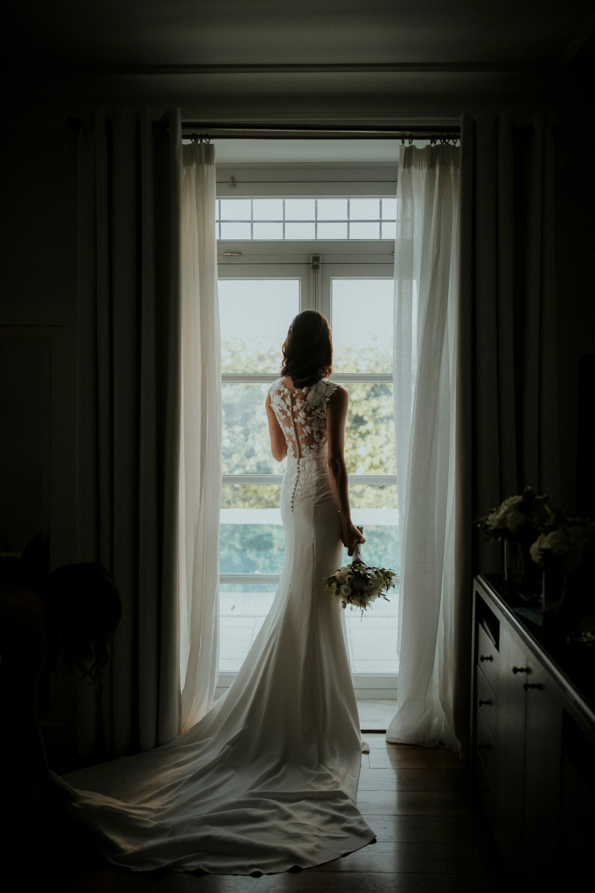 Bride in white dress looking out window holding bouquet