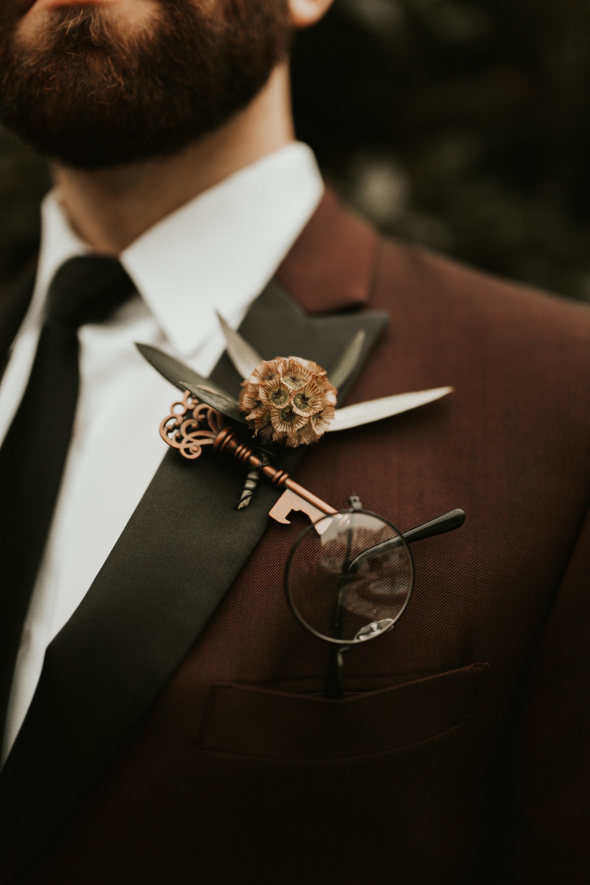 Groom in burgundy suit with quirky boutonniere and glasses