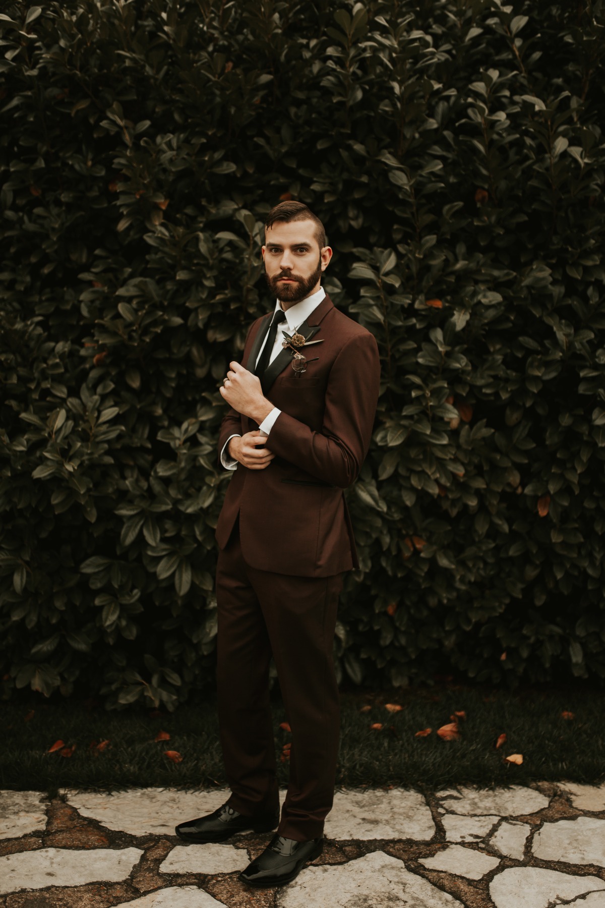 Groom in maroon suit posing before greenery backdrop