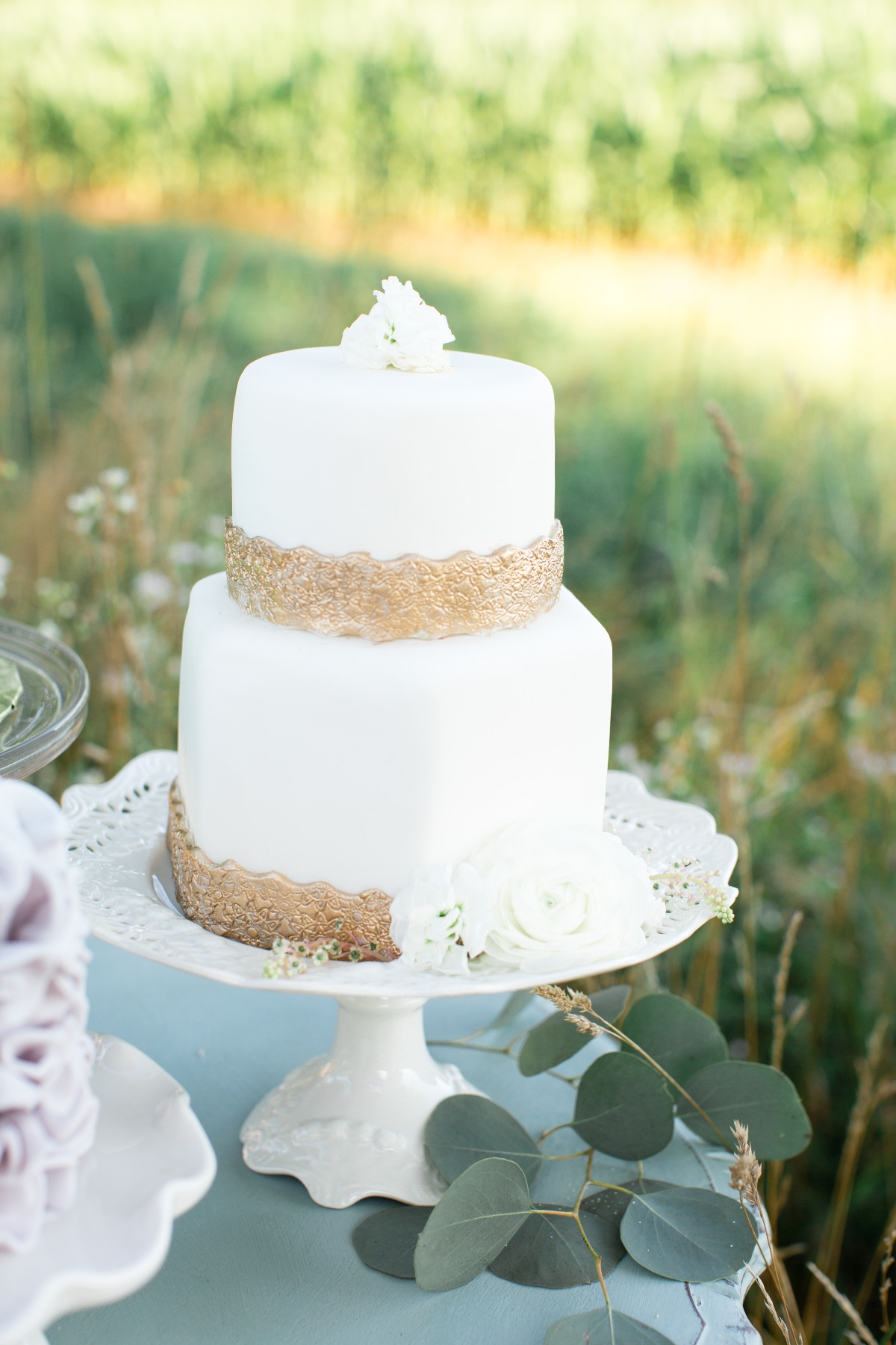 Elegant two-tier white wedding cake with gold detailing outdoors