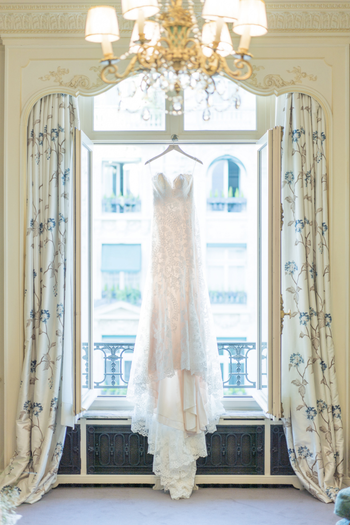 Lace wedding dress hangs in a window with chandelier